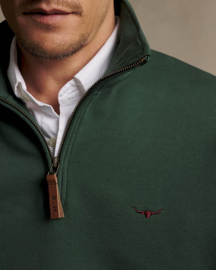 Close-up of a man wearing a dark green quarter-zip jumper with a brown leather zip pull and a small embroidered longhorn logo on the chest, layered over a white collared shirt.
