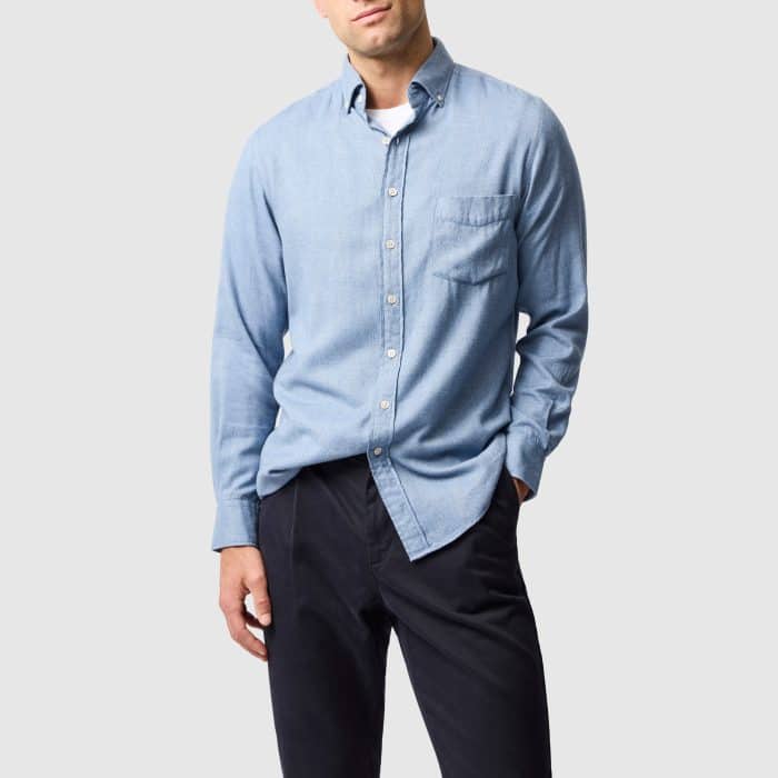 A man wearing a light blue button-down shirt over a white vest and dark trousers.