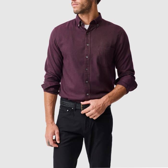 A man wearing a long-sleeved, button-through plum shirt with a breast pocket, paired with black trousers and a dark belt.