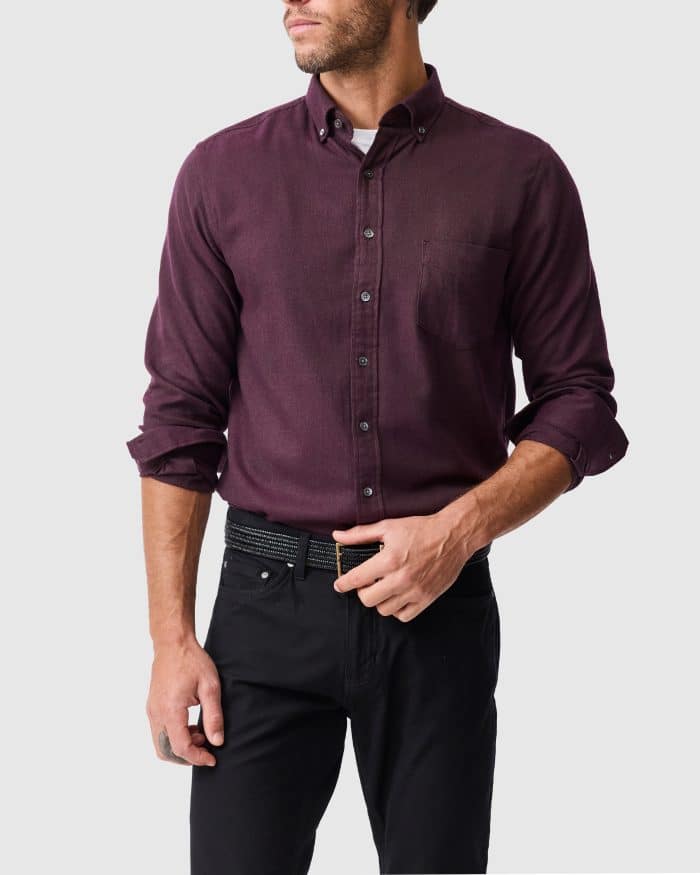 A man wearing a long-sleeved, button-through plum shirt with a breast pocket, paired with black trousers and a dark belt.