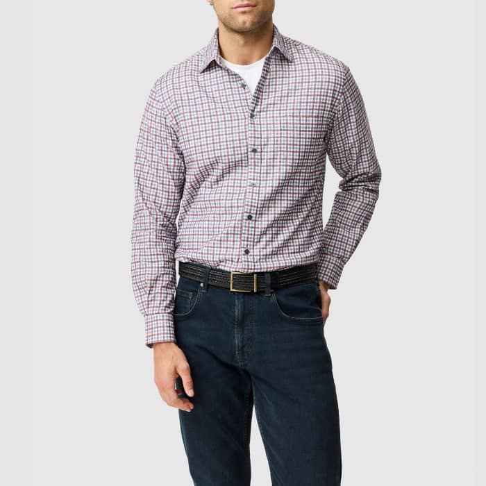 A man wearing a white vest, a red and blue checked button-down shirt, a black belt, and dark jeans.