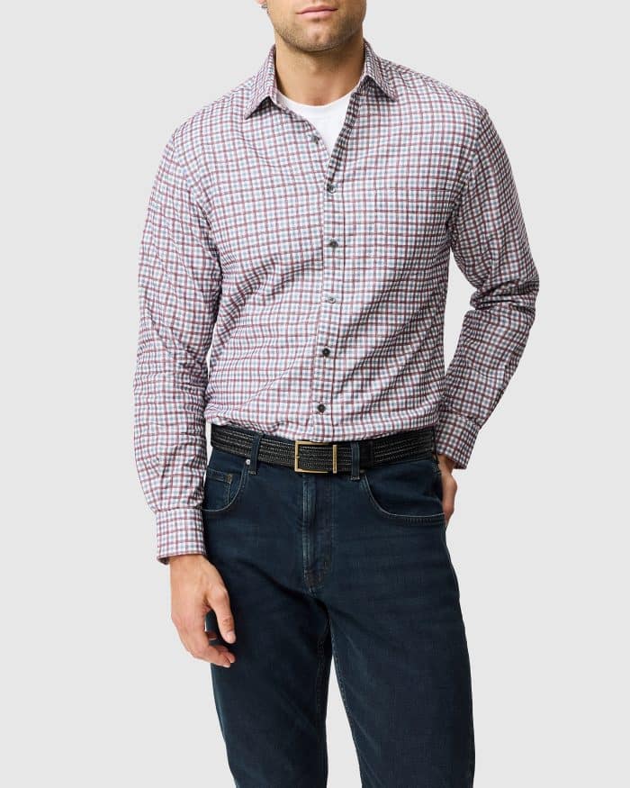 A man wearing a white vest, a red and blue checked button-down shirt, a black belt, and dark jeans.