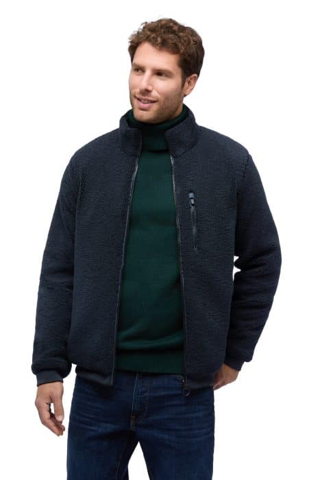 A man wearing a dark blue sherpa fleece jacket over a dark green polo neck jumper and blue jeans.