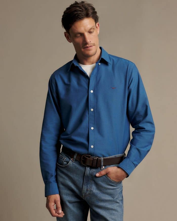A man wearing a blue button-down shirt over a white t-shirt, tucked into blue jeans with a brown leather belt.