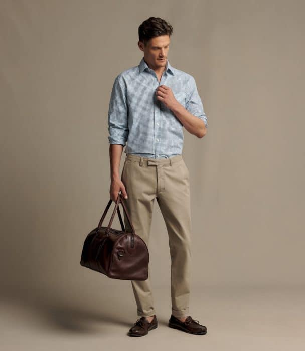 A man wearing a light blue checked shirt, tan trousers, and brown loafers, holding a dark brown leather holdall.