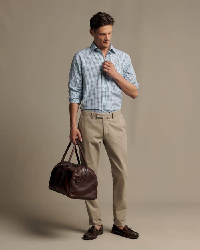 A man wearing a light blue checked shirt, tan trousers, and brown loafers, holding a dark brown leather holdall.